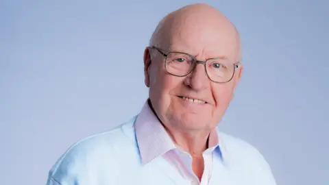 John Bennett wearing glasses and a pale blue jumper, pink shirt, smiling at the camera