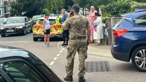 A soldier stands on the phone with his back to camera as people look on at the scene in Sally Port Gardens, Gillingham