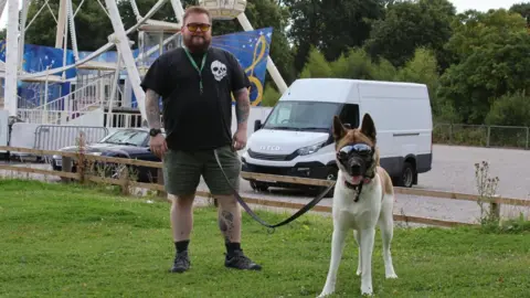 An akita dog wearing sunglasses stands by her owner on a lead. In the background, a car park, a van and a ferris wheel can be seen..