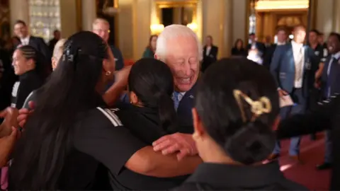 King Charles grins as three rugby players wearing black New Zealand t-shirts give him hug