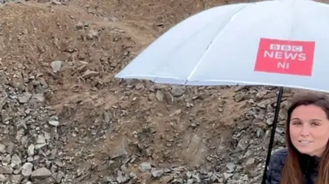 BBC reporter Faye Kidd standing with an umbrella in a rock-filled crater