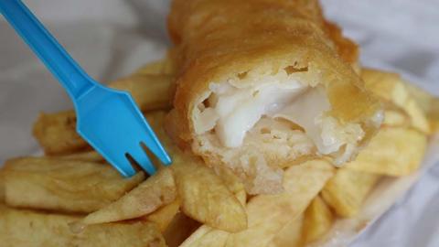 Close up of a classic portion of chip shop fish and chips. The fish is golden batter on the outside and white and flakey on the inside. A blue plastic fork is stuck in the chips