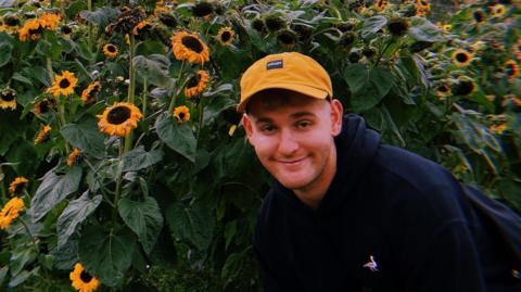 A man stands in front of a field of sunflowers wearing a yellow baseball cap with a blue hooded top and smiles at the camera