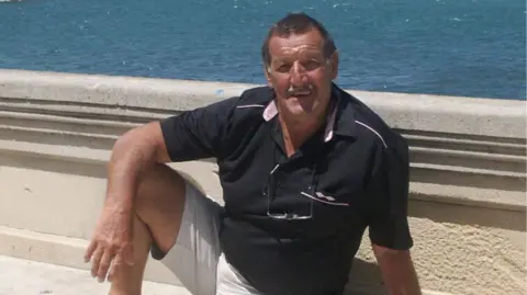 FAMILY PHOTO Former professional footballer Colin Flatt posing in front of a concrete seawall with the sea in the background. The image shows colin with a black polo shirt on and beige cargo shorts. He is sitting on the ground smiling, aged around 70.