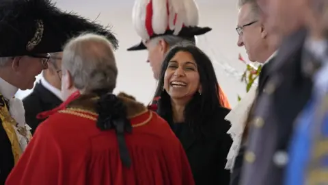 Kirsty Wigglesworth UK Home Secretary Suella Braverman laughs. She is surrounded by officials.