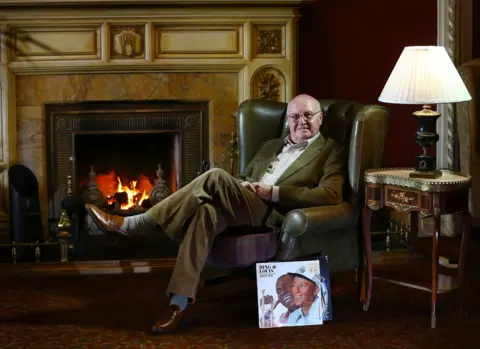 John Bennett sat in green arm chair with fireplace in background, table and lamp beside him and a record sat on the floor