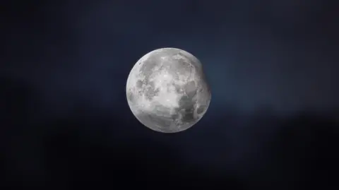 Gabriel The Moon is seen with a tiny part of the top-right covered by a partial eclipse. The Moon looks white in a clear sky.