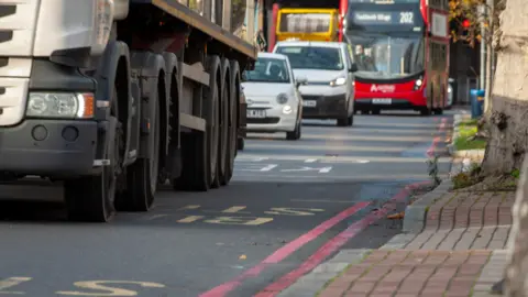 A close-up shot of the lower half of a lorry and then two white vehicles and a red bus behind it on a road in London