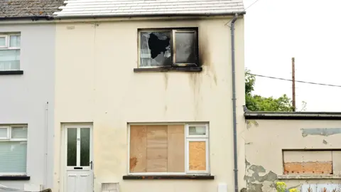 A cream coloured house with boarded windows and an upstairs window that is smashed and burned. There are burn marks down the wall.