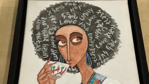 A framed drawing of a lady drinking from a cup, with words like "love", "hope" and "unity" written on her hair
