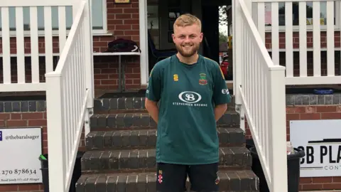 Sam Goodwin, wearing a green club shirt and smiling at the camera. He is standing in front of some brick stairs which have painted white wooden railings