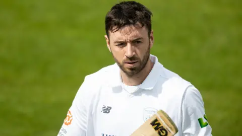 James Vince looks off camera while walking on the cricket pitch, he is wearing whites and carrying a cricket bat. He has dark brown short hair with a trimmed full beard.