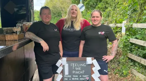 Three women stand in front of an advertising board with feeling the pinch written on it