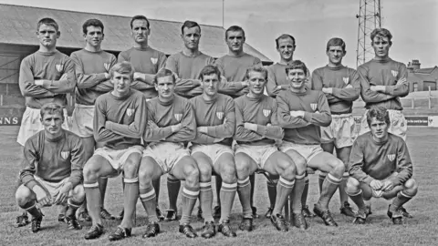 GETTY IMAGES A picture of the Leyton Orient League Division 2 team in 1965 in their full football kit. There are two rows of players in white shorts and long sleeved jerseys. The goalkeepers are crouched at either end. Colin Flatt is in the middle of the picture on the front row.