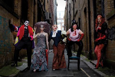 Five drag performers pose in an alley. The first, dressed as a man wears a white face and multi-coloured coat. The second has a grey dress with a large round hate. The third wears a blazer and wide trouers, the third stands on a chair in a t-shirt and red elbow-length gloves. The last rests with her back on the wall wearing red patterned tights and a corset.