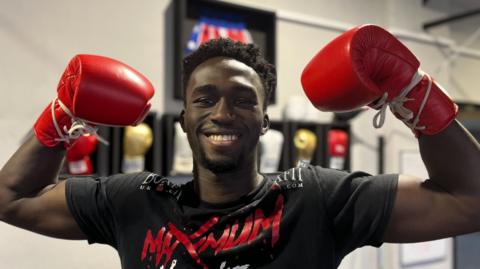 Adam Olaore smiles as he holds his arms up in a victory pose while wearing red boxing gloves