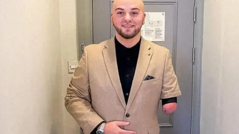 Ryan Rix who is bald with a beard and one forearm, wearing a light brown suit, standing in front of a grey door