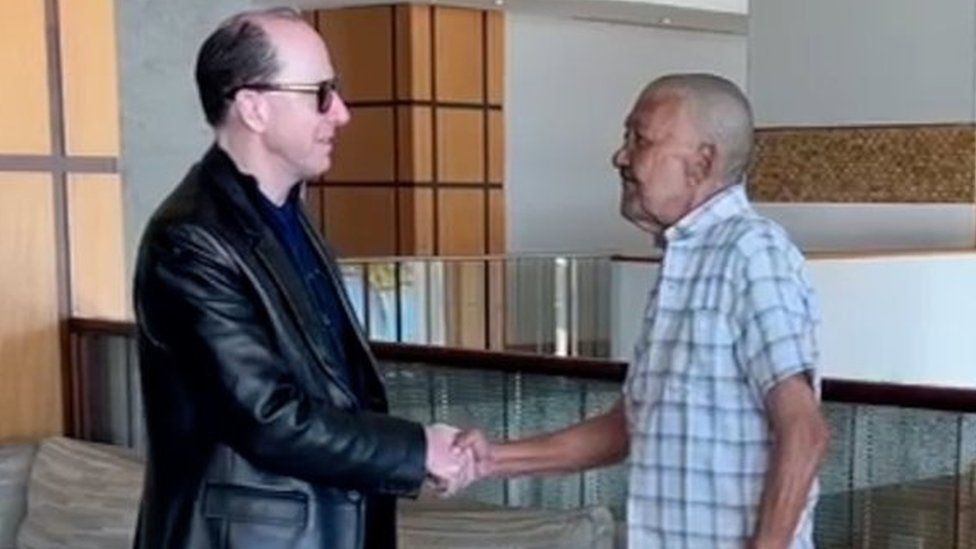 Mark Randolph Dyer (left) is dressed all in black. He is shaking hands with Nizamodeen Hosein