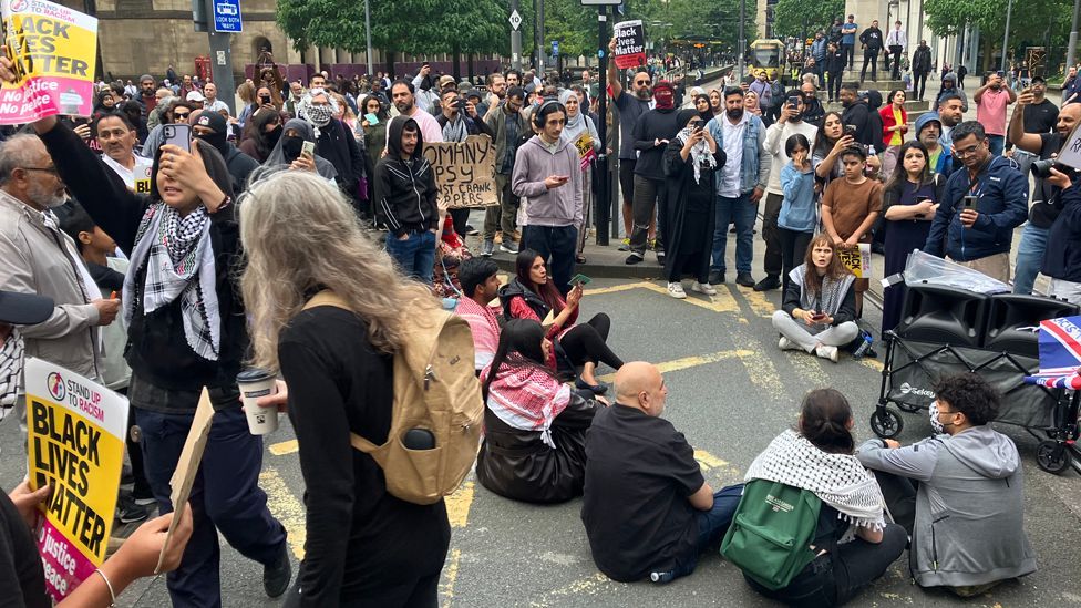 A number of protesters holding signs and standing and sitting in the road during a protest in Manchester