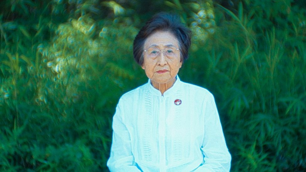 Portrait of Michiko Kodama, an elderly woman with short dark hair. She is wearing metal-rimmed glasses and has a serious expression. She is pictured standing in front of a green bush.