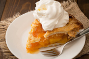 A high angle extreme close up horizontal shot of a slice of freshly baked peach pie revealing the golden fruit inside. The flaky crust is topped with a generous portion of whipped cream. The pastry and metal fork sit on a white plate which along with the cream provide some  copy space for your message