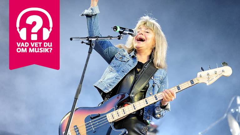 Suzi Quatro har en elbas kring halsen, höger arm i luften och sjunger i en mikrofon.