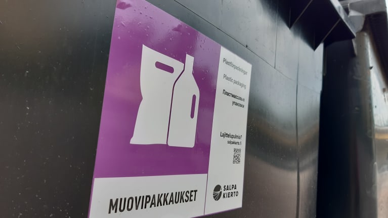 Mustan muovisen astian kylki, johon on liimattu lilan ja valkoisen värinen tarra. Tarrassa on symbolinen kuva pussista ja pullosta ja alla lukee muovipakkaukset.