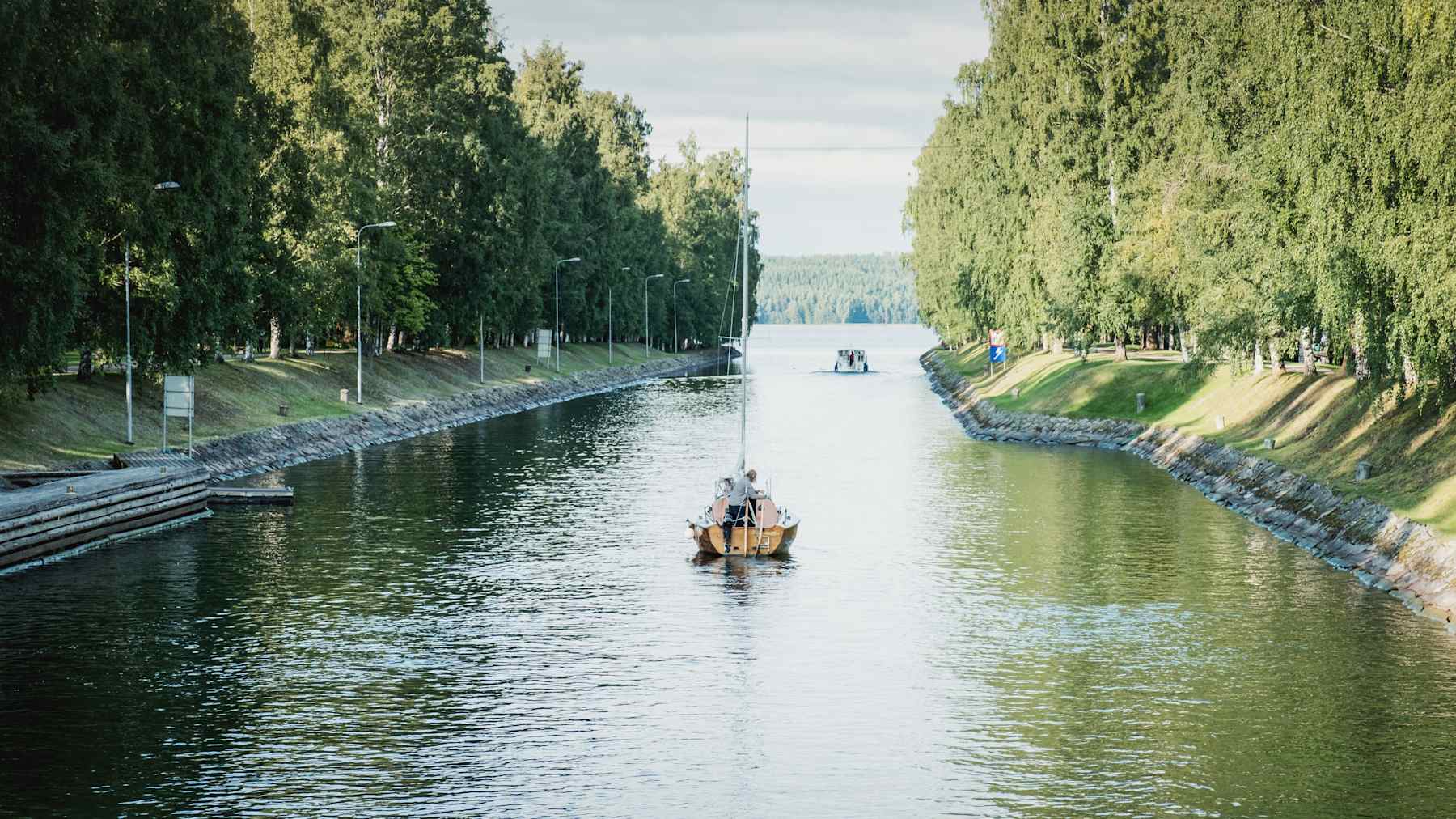 Segelbåt på väg bortåt längsmed kanal, längre bort skymtas mynningen till en sjö. 