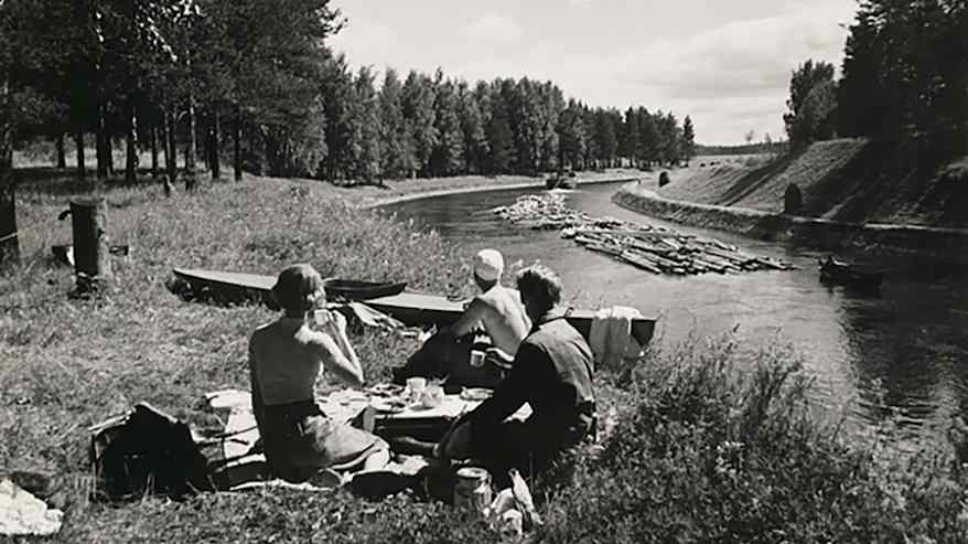 Svartvit bild: Tre personer har piknik vid kanalstrand, stockar flottas genom kanalen.