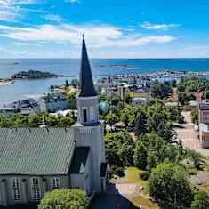 En bild tagen högt upp över en stad vid havet. I förgrunden en kyrka och gröna träd. I övrigt bostadhus varvade med ämbetshus och gröna träd. Längs bort ses klippor och skär och ett blått hav som möter en blå himmel med små molnslöjor här och där. Det är grönt och lummigt på bilden.