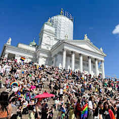 Crowd of colourfully dressed people on the steps of a large white cathedral in sunny weather.