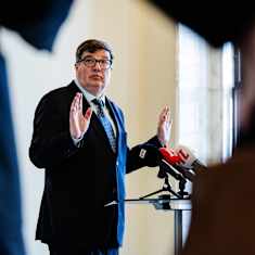 A man in a dark blue suit and glasses stands in semi-profile with his hands partially raised behind microphones on a podium.