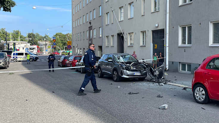 Poliiseja kadulla, puoliksi tuhoutunut auto ja katselijoita kadulla.