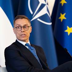 Alexander Stubb sits in an armchair on stage wearing a suit. The EU, NATO and Finnish flags are in the background.