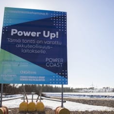 A large blue sign saying Power Up! and Power Coast with CNGR and Hamina logos stands in a partly snowy field with wind turbines in the distance.