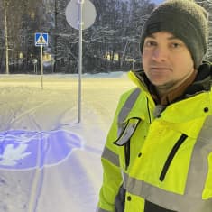 En man i gul reflexjacka och mössa står på en snöig cykelväg med ett upplyst trafikmärke på marken. 