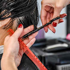 Närbild på två händer som håller på att klippa en person med mörkt kort hår.