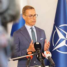 A man in a blue suit and brown horn-rimmed glasses speaks with a serious expression at a podium with microphones; Finnish and Nato flags behind him.