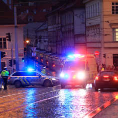 En ambulans och en polisbil står vid ett gathörn. Blåljusen speglas i den våta stenläggningen på gatan.