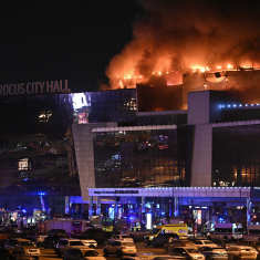 Smoke rises from a large building with a sign reading 'Crocus City Hall' at night, with many emergency vehicles and cars in front of it.