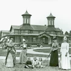 Fyra kvinnor i långa klänningar och fina hattar framför en utsmyckad träbyggnad. Mellan dem sitter tre barn i gräset. Kvinnorna håller alla i tennisracketar. Bilden är svartvit och från 1910.