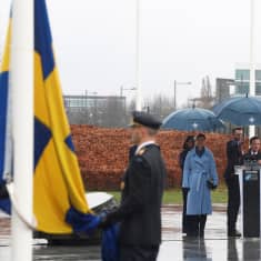 Sveriges flagga hissas vid Natos högkvarter i regnväder. Kronprinsessan Victoria, statsminister Ulf Kristersson och Natos generalsekreterare Jens Stoltenberg ser på. Bakom dem står tre assistenter och håller paraplyer över dem.