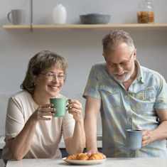 medelålders man och kvinna står i ett kök med kaffemugar i händerna, båda skrattar.