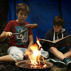Två pojkar sitter i ett vindskydd och läser/grillar
