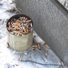 En stor konservburk full med tobaksfimpar står vid en vägg. På marken syns snö, fimpar och tändstickor.
