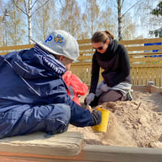 En kvinna gräver med liten spade i sandlåda. Framför henne till vänster syns en ung pojke bakifrån, han gräver också i lådan.