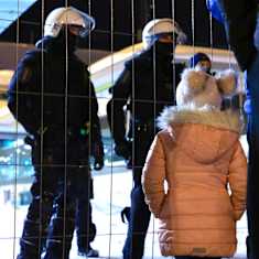 Two Border Guard officers wearing helmets facing a child wearing winter clothes behind a fence at the Vaalimaa border crossing.
