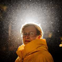 Ulla-Maj Wideroos i en gul rock tittar mot kameran utomhus då det regnar. Det är mörkt ute.