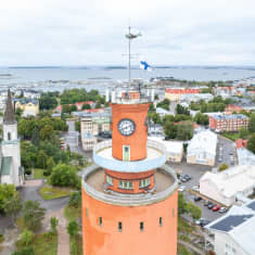 Ett rostrött torn med mintgröna detaljer. Omkring tornet växer träd och i bakgrunden syns trähus och mindre betongbyggnader. Längre bort syns havet