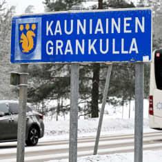 En skylt där det står "Kauniainen - Grankulla" vid en väg. På vägen kör en grå personbil och en buss. Det är snöigt på vägen.